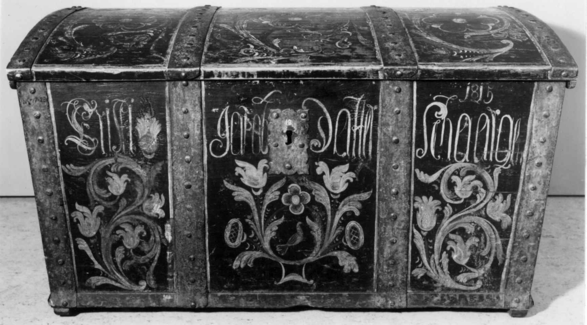 Kiste med buet lokk og smijernsbeslag og bånd. Rosemalt lokk og tre sider. innvnedig bemalt og med leddik. Kisten har små føtter.