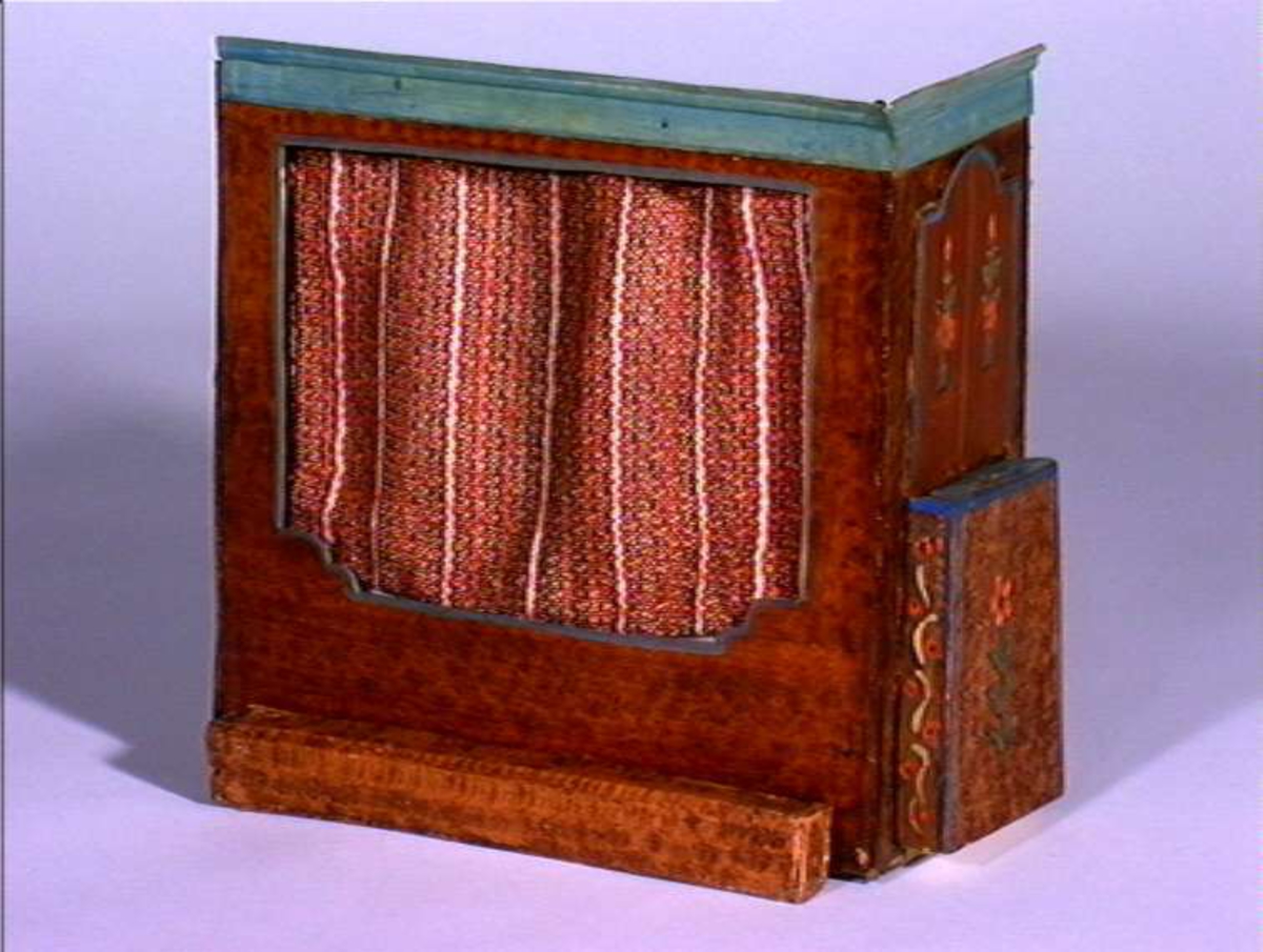 Dukkestue i form av svensk hytte med påmalte tømmerstokker, grunnmur og takstein i tegel. En vegg kan løftes bort og taket kan også åpnes. Stuen har tre stk vinduer med påmalte blysprosser. Inne i stuen er peis, bord og en dukke, en man i svensk folkedrakt med fele, fastmontert. På veggen henger et kurbitsdekorert maleri av Carl Johan.