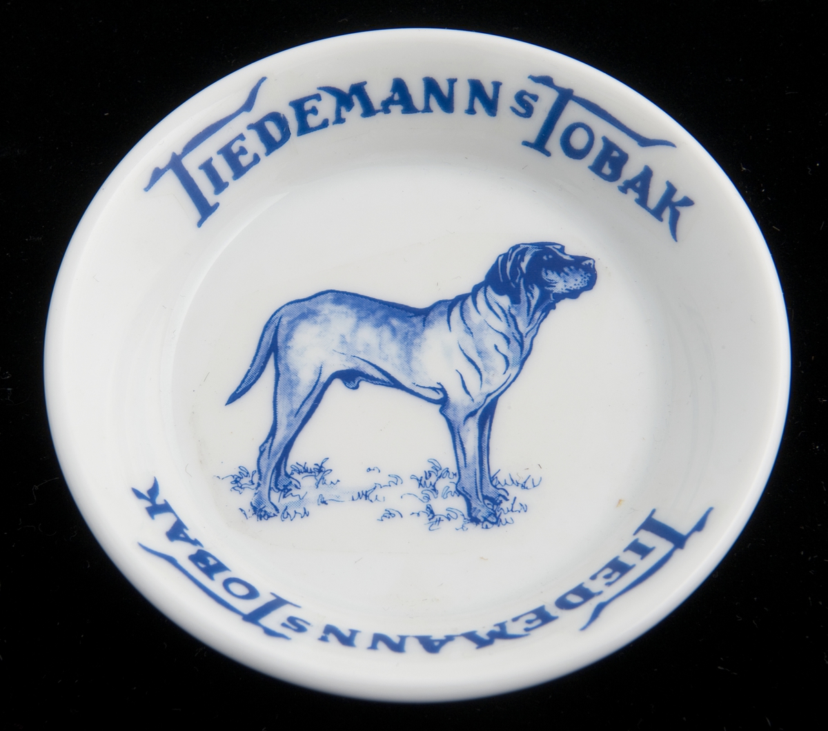 Liten, rund skål i porselen med reklame for Tiedemanns Tobak. Påtrykket motiv i blått av en stående hund.
