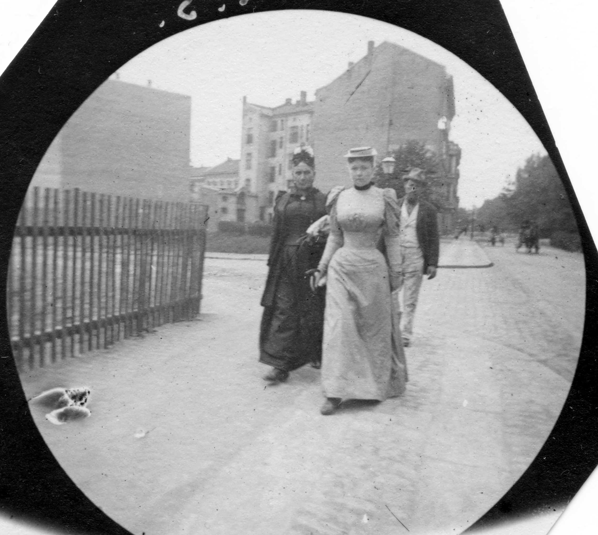 Frk. Florens Sinclair og hennes mor, fru Sinclair spaserer langs gjerde med bygårder i bakgrunnen, Oslo.