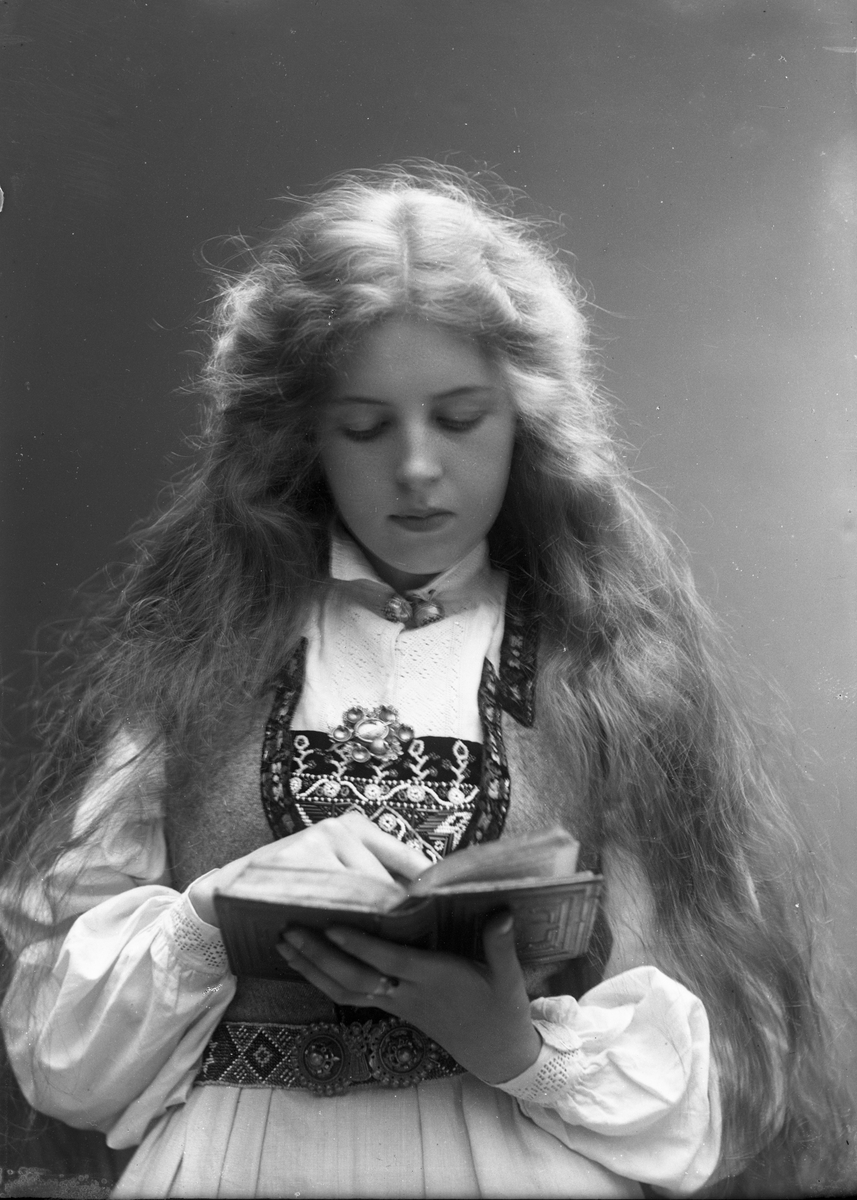 1/2 dels studioportrett av kvinne i drakt med utslått hår. Hun holder en bok i hendene som hun leser i. Bakgrunnen er nøytral.