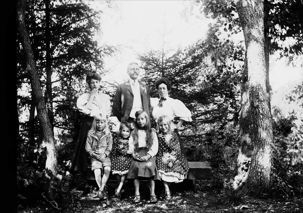 Familiegruppe, fire barn sitter på benk, en mann og to kvinner står bak, ukjent sted.