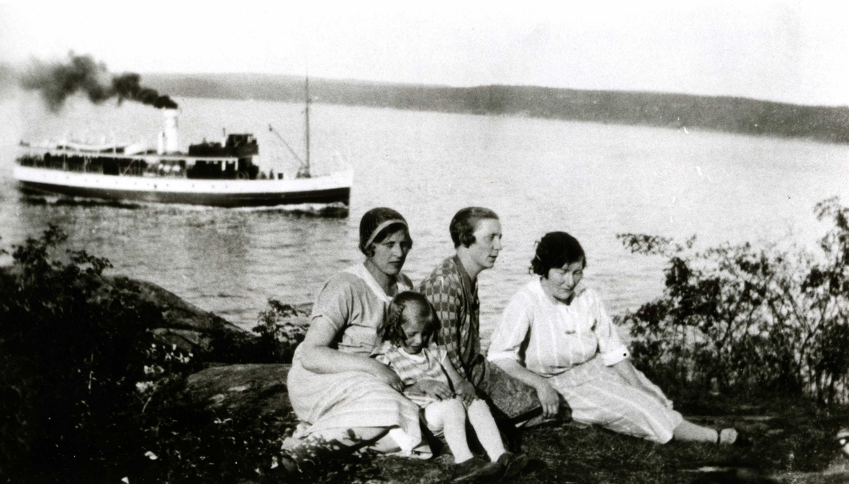 To kvinner, en mann og et barn sitter på bakken med ryggen mot sjøen. I bakgrunnen Nesoddbåten på vei innover Bunnefjordsen. Ferieliv på Oslo Godtemplarungdomslags feriehjem Kirkevik, Nesodden