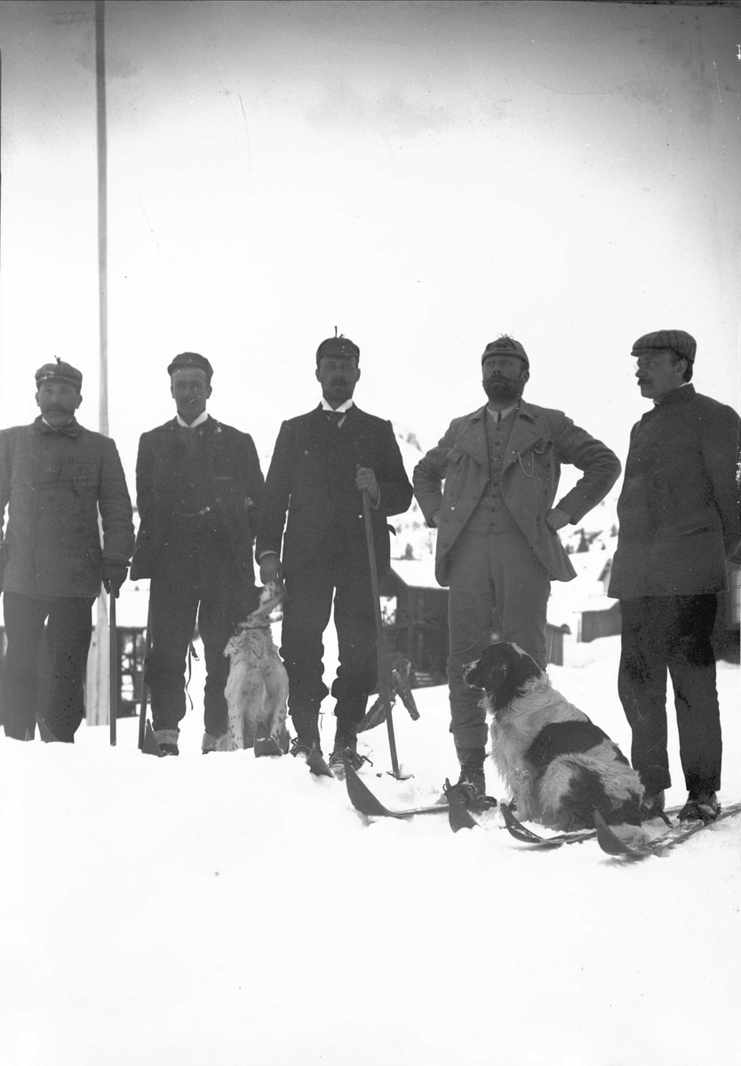 Fem menn ute i snøen, to sittende hunder, flaggstang og bygninger i bakgrunnen. Muligens Fefor sanatorium, Nord-Fron i Gudbrandsdalen.