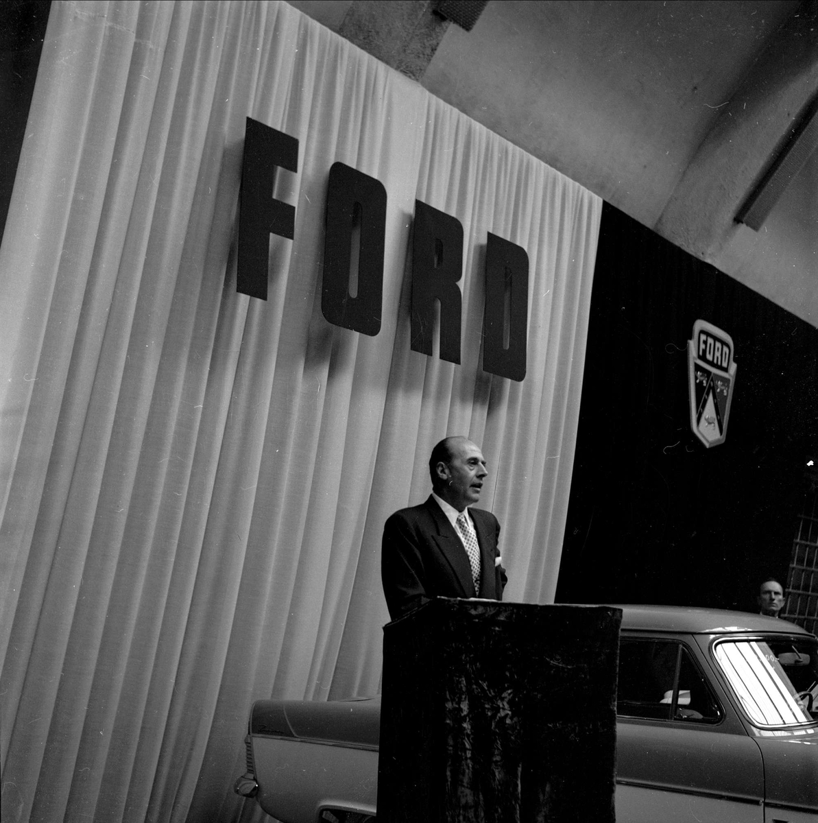 Bilutstilling, merke Ford, antakelig Oslo, 10.09.1956. Mann taler, kanskje nybilpresentasjon.