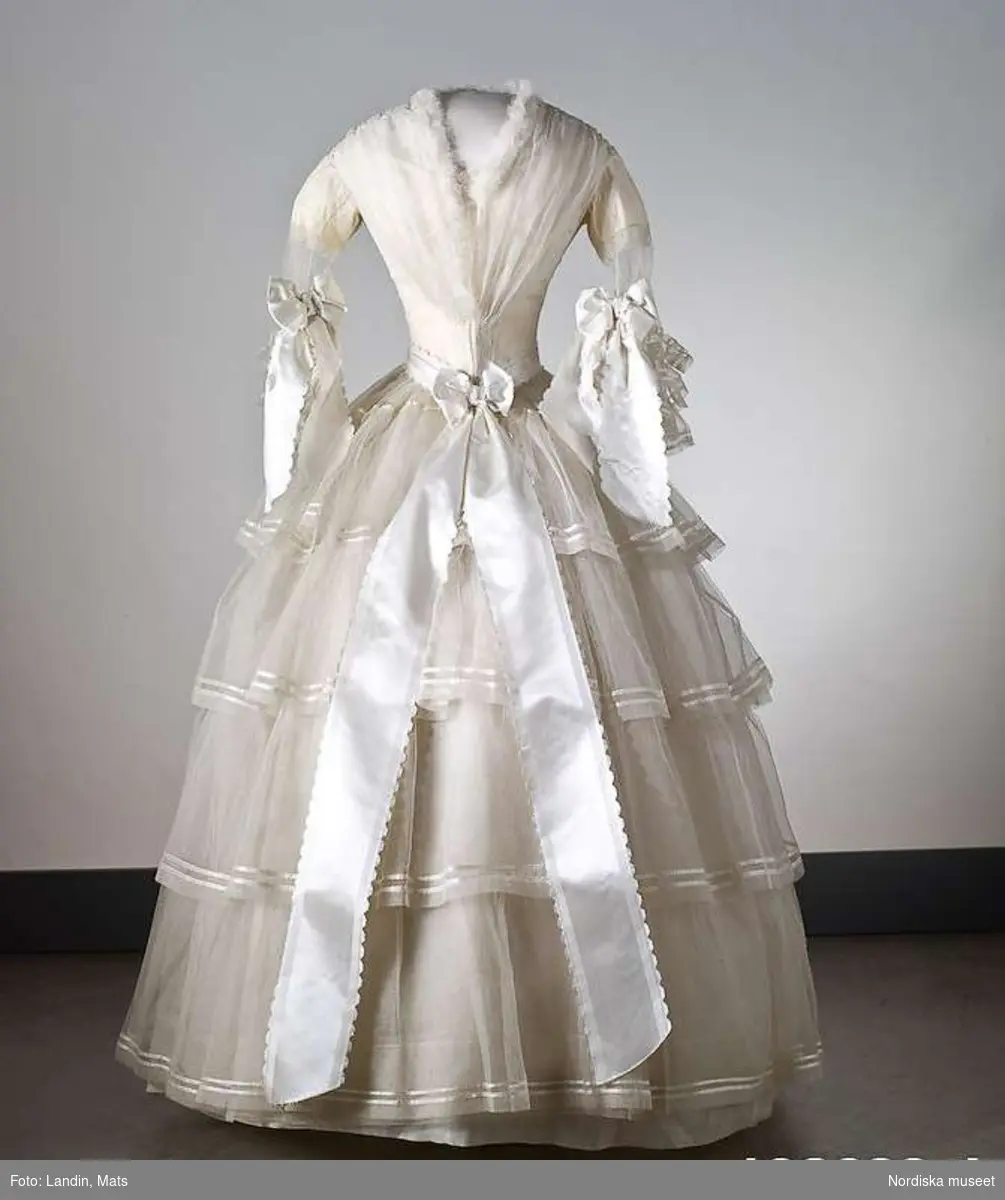 Brudklänning, liv med fastsydd kjol, vit tarlatan, garnerad med vita sidenband, rosetter. Klänningen har ett löst skärp av vit tarlatan.