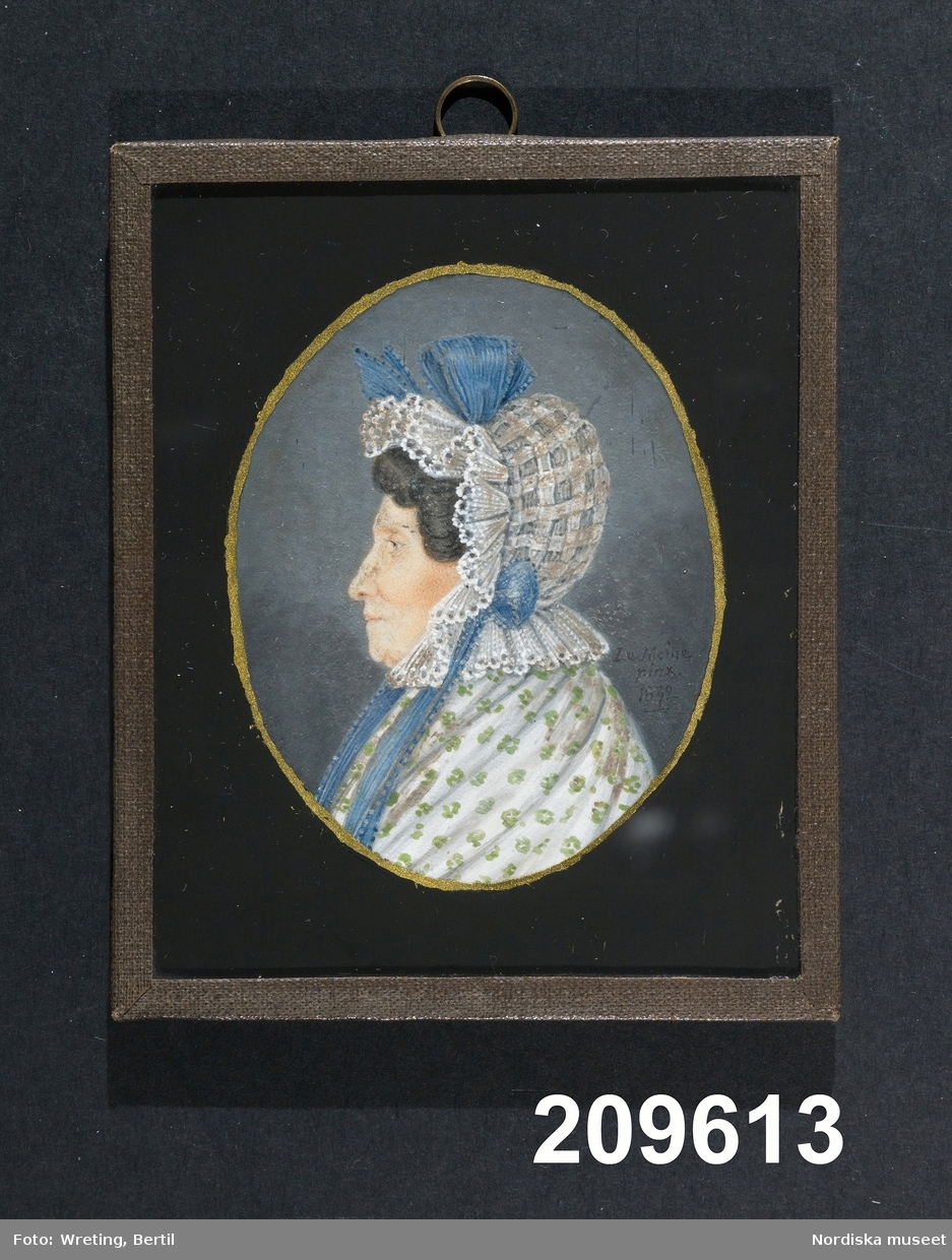 Profil, bröstbild. Mörkhårig kvinna med rutig spetsprydd mössa, blå rosett och blåa band. Vit sjal med grönt mönster. 
/Annika Tyrfelt 2015