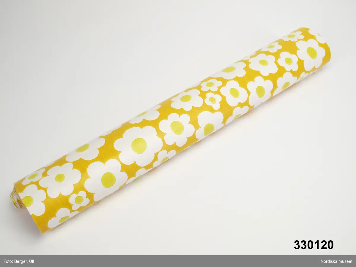 Tapetprov, del av tapetrulle. papper, tryckt mönster, enkla vita blommor med gul mitt mot gul botten. Inga stämplar på baksidan. Kantskuren.
/Leif Wallin 2010-07-09