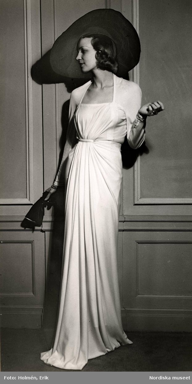 Dammode, kvinnlig modell poserar iklädd draperad aftonklänning och vidbrättad hatt. I handen håller hon ett par handskar. Nordiska Kompaniet 1938.