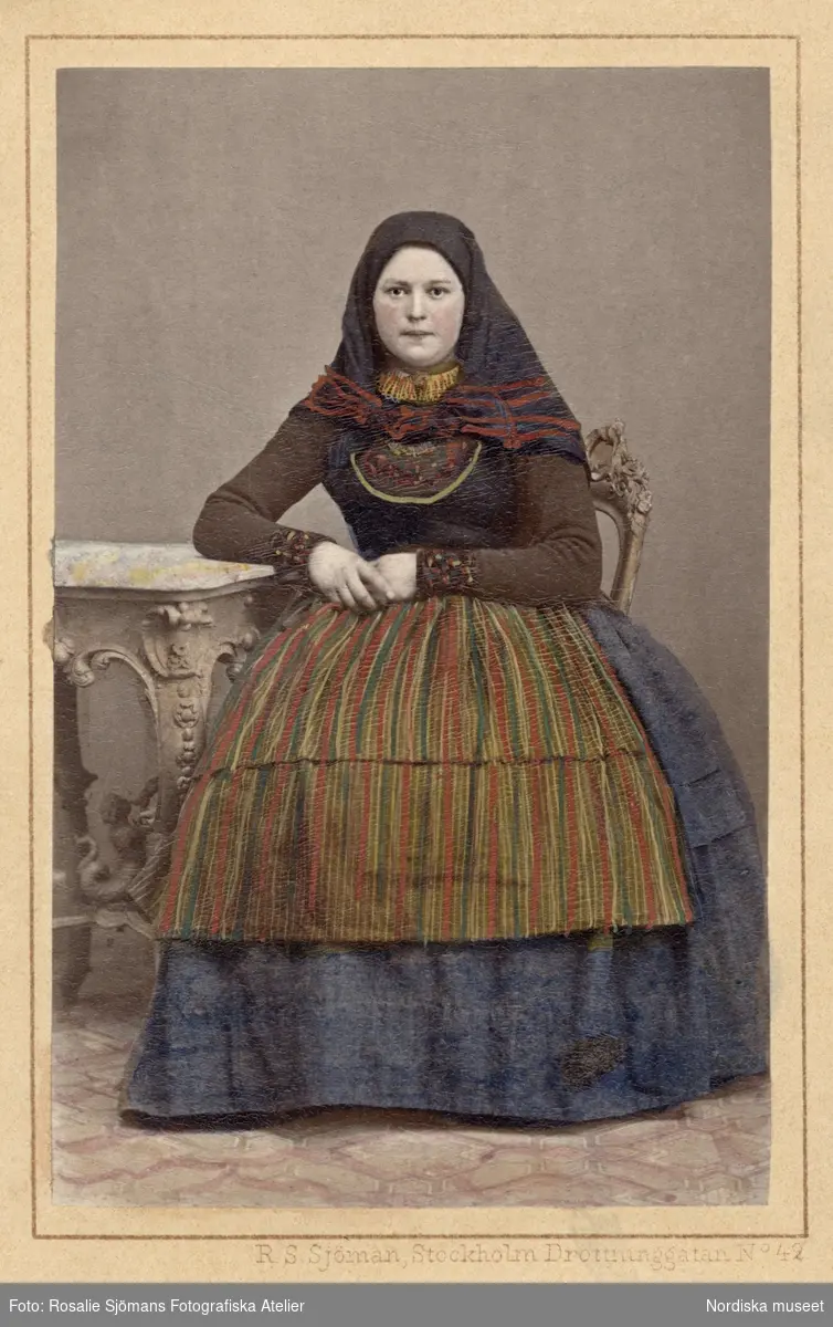 En kvinna i folkdräkt poserar sittande iklädd sjalett och randigt förkläde. Genarp, Skåne. Kolorerat i efterhand. Nordiska museet iv.nr. 18536C.