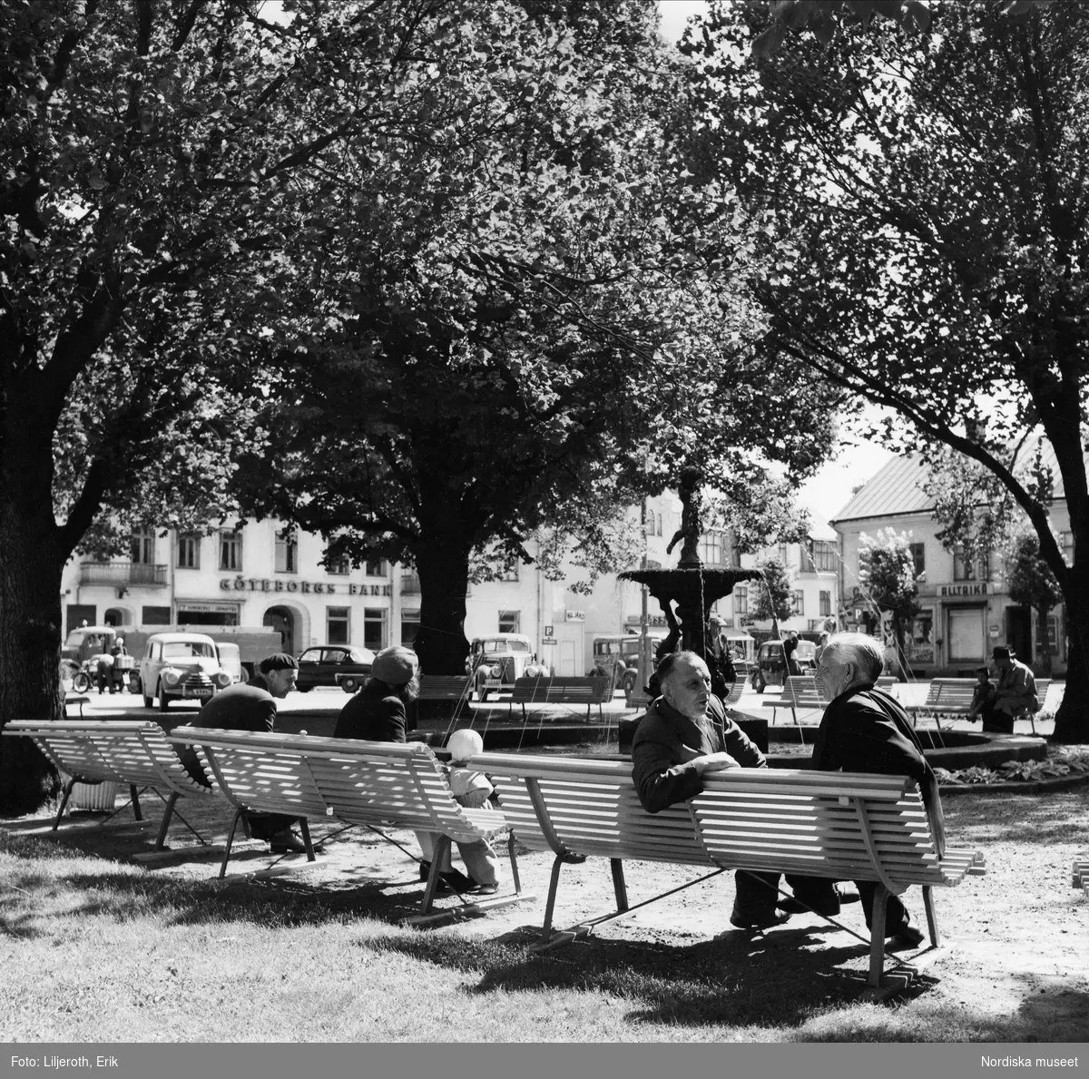 Stortorget i Nässjö. På parkbänken närmast några män inbegripna i samtal. Vuxna och barn i solen.
