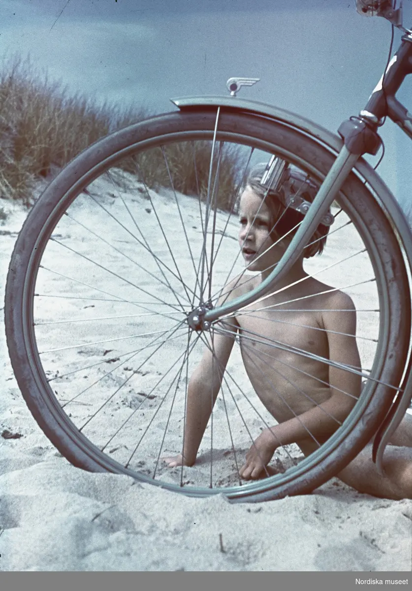 Flicka på sandstrand. Hon tittar fram genom ekrarna på ett cykelhjul.