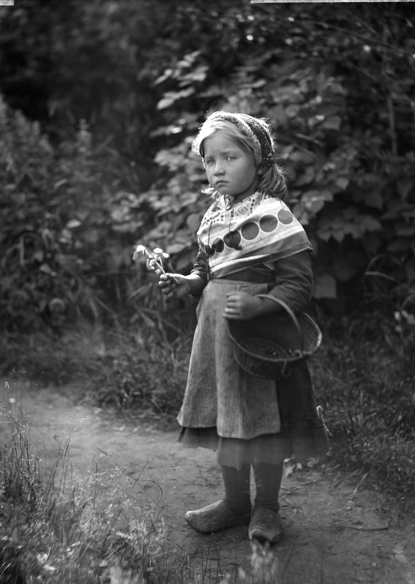Liten flicka med korg och blommor i händerna. Flickan är Selma Andersson (född 1893), dotter till en torpare. Hon finns på många av Severin Nilsons foton och tavlor. Schalen är Severin Nilsons rekvisita och återfinns också den på flera andra foton. 