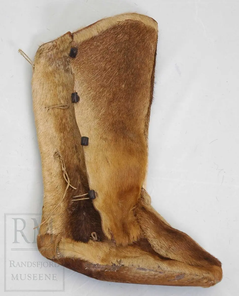 Høye støvler i skinn med pels på ytterside. Såle, overlær og skaft av samme materiale. Skaftet har åpning i siden og lukning med knapper og hemper. 
