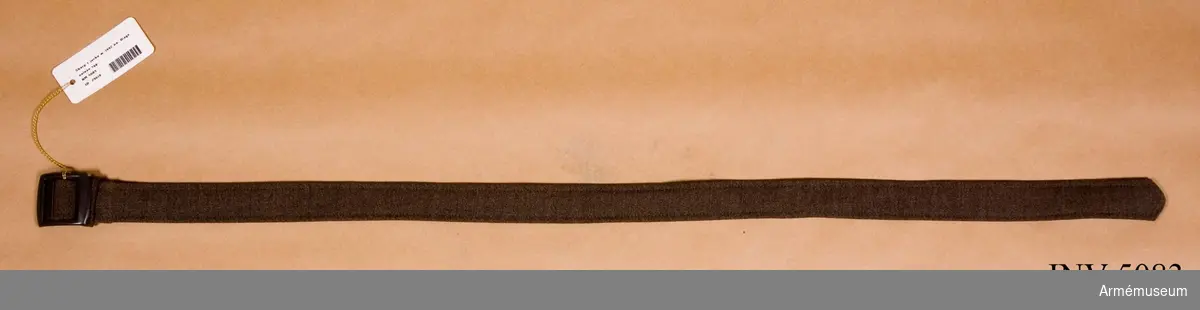 Diagonalväv tyg. Skärpet fastsättes i härför avsedda hällor på jackan i sidorna. Skärpet har sölja av samma tyg och ett spänne i mattgrå färg.