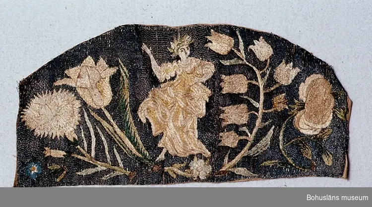 Broderi på beige linne, med rak nederkant och rundad överkant. 1700-talet.
Motivet är en dansande figur med bockfot (kvinna?) omgiven av blommor (en nejlika, en tulpan, en klockhyacint?, en blå blomma och en ros).
Mönstret är gjort av silke i schattérsöm. Himlen i bakgrunden är sydd av metallöverdragen silketråd (troligen silver) i läggsöm.
Färgerna i broderiet är (på baksidan) olika blå nyanser, olika gröna nyanser, ljusgult, beige, rödtonat beige och brunt.

Ett par små hål i kanten. Brunt längs kanterna. Läggsömmen över en del metalltrådar är borta. Mycket blekt.

Knut Adrian Anderssons katalog:
1.26:14
Stycke av ett broderi från 1700-talet gjort av silke o metalltråd å grå botten. (31 x 16 cm.) Sk. av Göteborgs museum 3/4 1880 genom intendent A. W. Malm.

Ur handskrivna katalogen 1957-1958:
"Stycke av broderi. Nr 70, 71, 73 hör ihop.
1870
Mått: 31,5 x 15,5 cm; broderi som nr 70 - 71; dansande kvinna omgiven av blommor; ngt defekt."