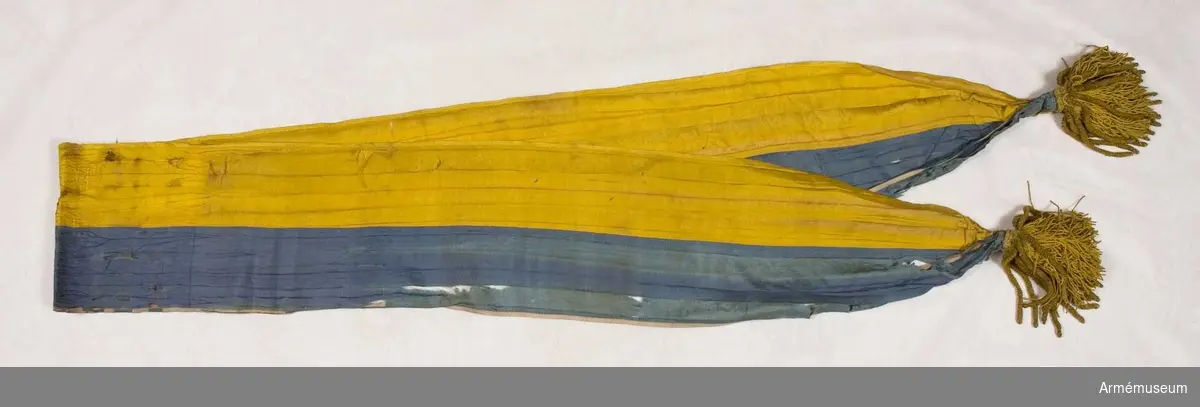 Kravatten (fanbandet) är på längden hälften blått och hälften gult. I vardera änden en tofs med i mitten gult tvinnat silke och utanpå detta grova guldbuljoner.