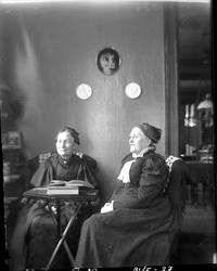 To eldre kvinner kledd i sort leser ved et lite bord