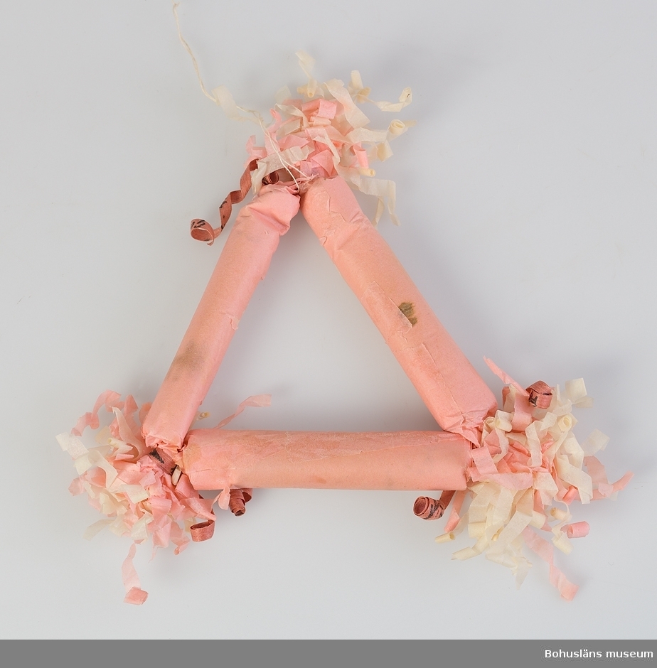Julgranskaramell, tre karameller lindade med rosa och vitt "krusat" silkepapper är hopknutna till en triangel.
Prydnad avsedd för julgran.
Clara Nilsson (1866-1946) var  mormor till givarna.