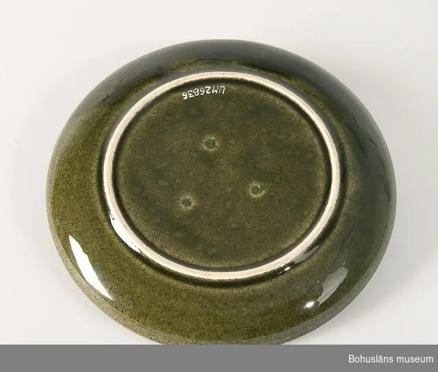 Assiett av keramik glaserad i melerat mörkt gröngult med brunt inslag. Slät, uppböjd kant ytterst.
Hör samman med kaffekopp UM26835, gräddkanna UM26837 och sockerskål UM268388. Ytterligare uppgifter se UM28835.