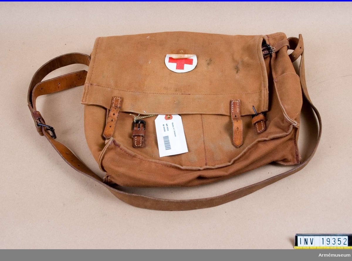 Nr 4. Bärbar väska med axelrem och spännremmar i läder och tillhörande spänne. Väskan är tillverkad av väv och läder och är märkt med  Röda Korsets märke.   