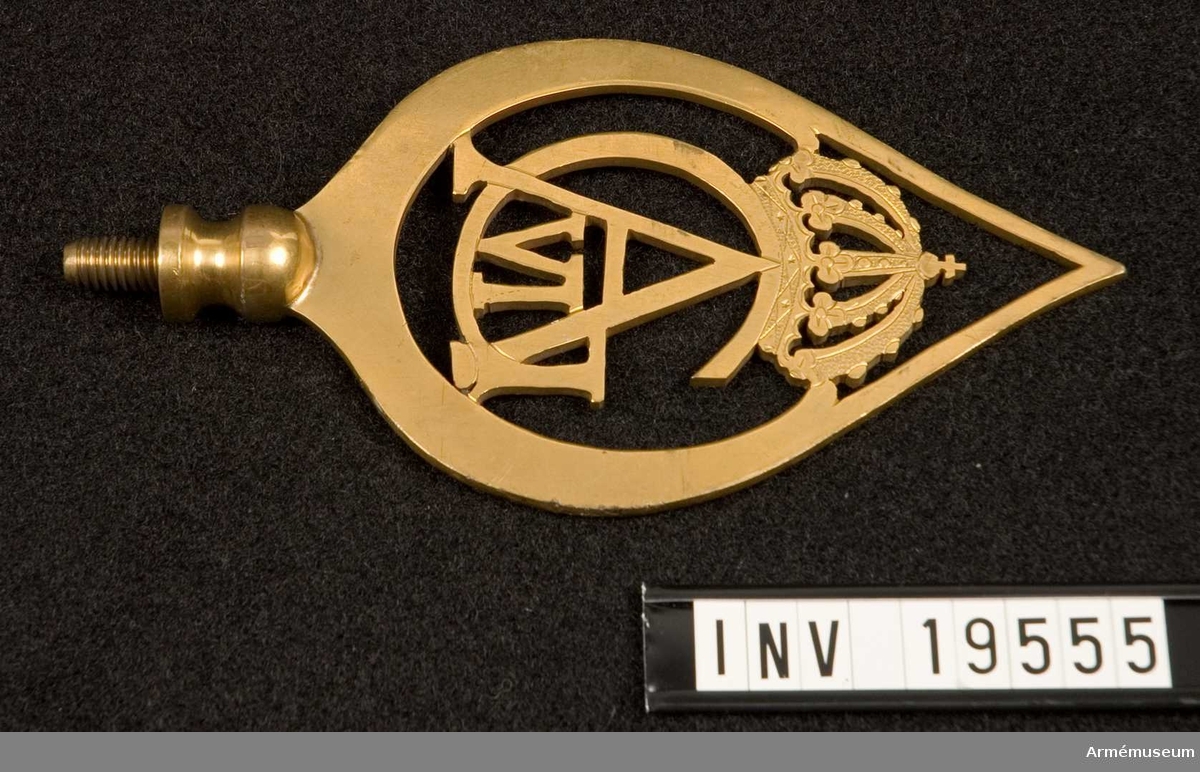 Spets till fana med Gustav VI Adolf:s monogram. Spetsen är tillverkad av förgylld mässing. Den har Gustav VI:s spegelmonogram under en kunglig krona, inramat av ett bladverk som avslutas i en spets. Spetsen är försedd med skruv.
