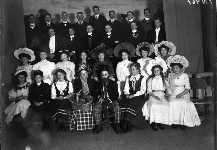 Enligt tidigare noteringar: "Gruppfoto av herrar och delvis utklädda damer uppställda på enkel scen. Kostymfest. Hantverksföreningen i Uddevalla."