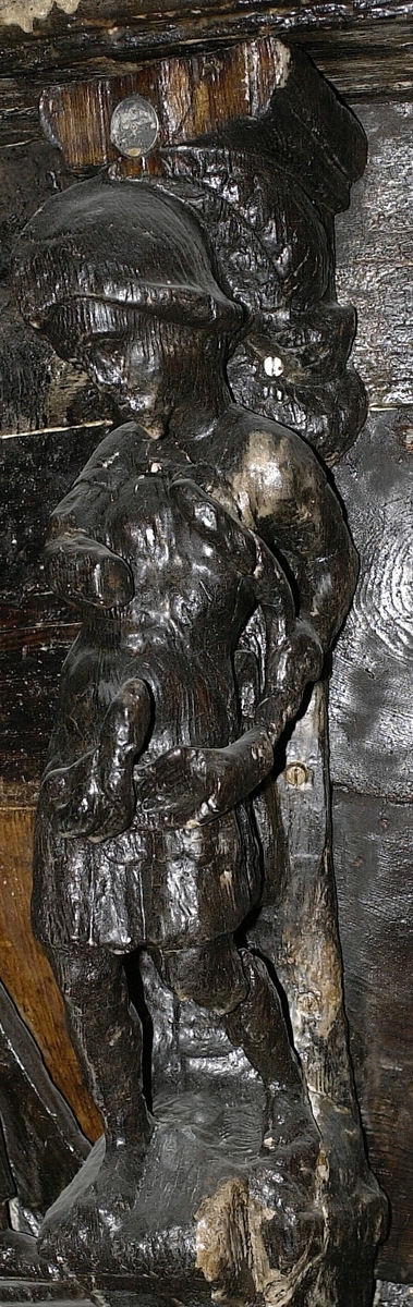 Krigare iklädd romersk rustning och hjälm med stora plymer. Huvudet är nedböjt och lutat åt höger. Rustningen är alltför sliten för att tillåta någon noggrannare analys. Mannen blåser i en stor, S-svängd lur som han håller i vänster hand. Med höger hand håller han en, nu fragmentarisk, fackla framför sig. Mannen står i en knäande ställning och huvudparten av kroppstyngden förefaller vila på höger ben. Sockeln visar svaga spår av profilering och "mark". Framsidan av sockeln är svagt triangelformig med spetsen nedåt.
Skulpturen är kraftigt sliten.

Text in English: Warrior, clad in Roman armour, clean-shaven and wearing a helmet with large plume. The head is bowed slightly and leaning to the right. The man blows a large, S-shaped horn held in his right hand. In his left hand he holds the fragments of a torch. The figure''s knees are slightly bent and the main weight of the body appears to rest on the right foot. The base shows small traces of profiling and supposedly depicts a clump of "earth". The front side of the base is vaguely triangular in shape.
The sculpture is badly worn.