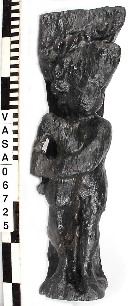 Skulptur föreställande en barnfigur spelande på en säckpipa.
Huvudet är vridet åt vänster och något nerböjt. Säckpipan hålls med bägge händerna och stöder dessutom på höger ben som är böjt. Kroppstyngden vilar på vänster ben.
Ovanför hjässan ses en snedställd äggstavslist. Det triangelformade partiet ovanför listen har utgjort anliggningsyta. Figuren står på ett minimalt underlag som troligen avser att föreställa ett stycke naturlig mark. Skulpturen har en slät baksida.
Skulpturen är kraftigt sliten och ytnött.

Text in English: This is a sculpture of a child playing a set of bagpipes.
The bagpipes are held in both hands and are further supported on the right leg, which is bent. The left leg is straight and takes the weight of the body. The head is turned slightly to the right and a trifle bent. The figure is partly draped in a cloak which conceals his right side.
Above the head the backboard comes forward at right angles. The front of the board is wide and is decorated with an egg-moulding design  which is placed diagonally across it, with the left-hand side lower than the right hand side when viewed from the front.. The figure stands upon a small flat plinth. The back of the sculpture is smooth.
The sculpture is badly worn.