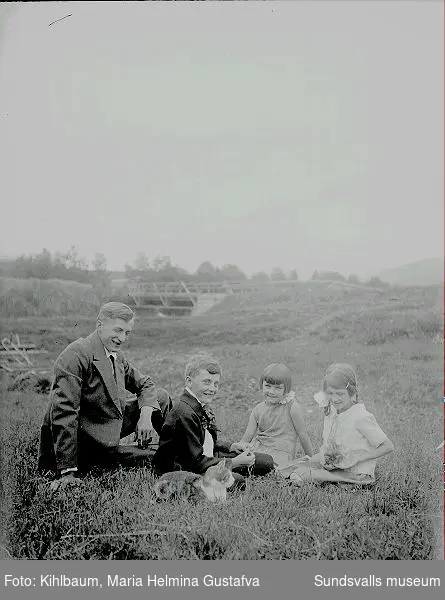 Grupporträtt. Man, en pojke och två flickor plus en katt sitter i gräset. I bakgrunden en bro.