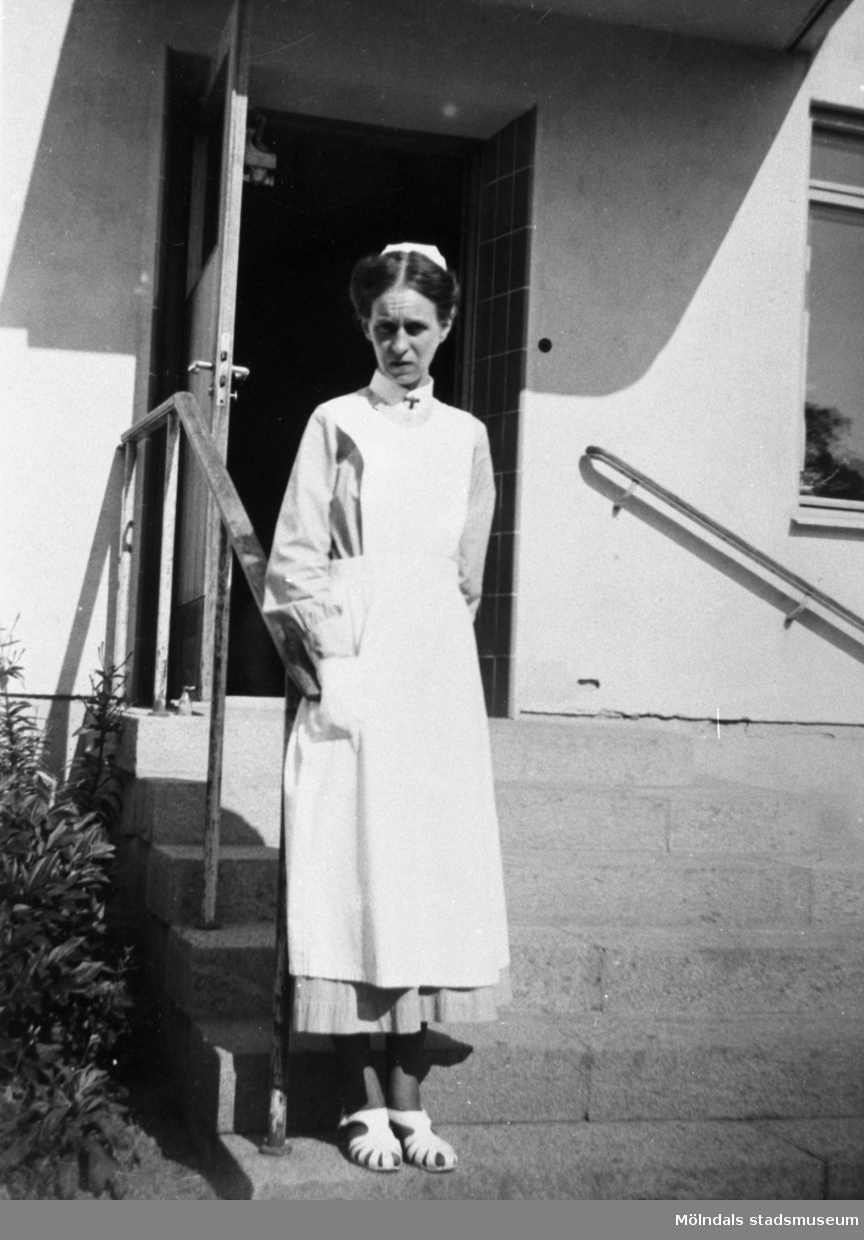 Kvinna, troligen sköterska, stående på trappan utanför ett hus.