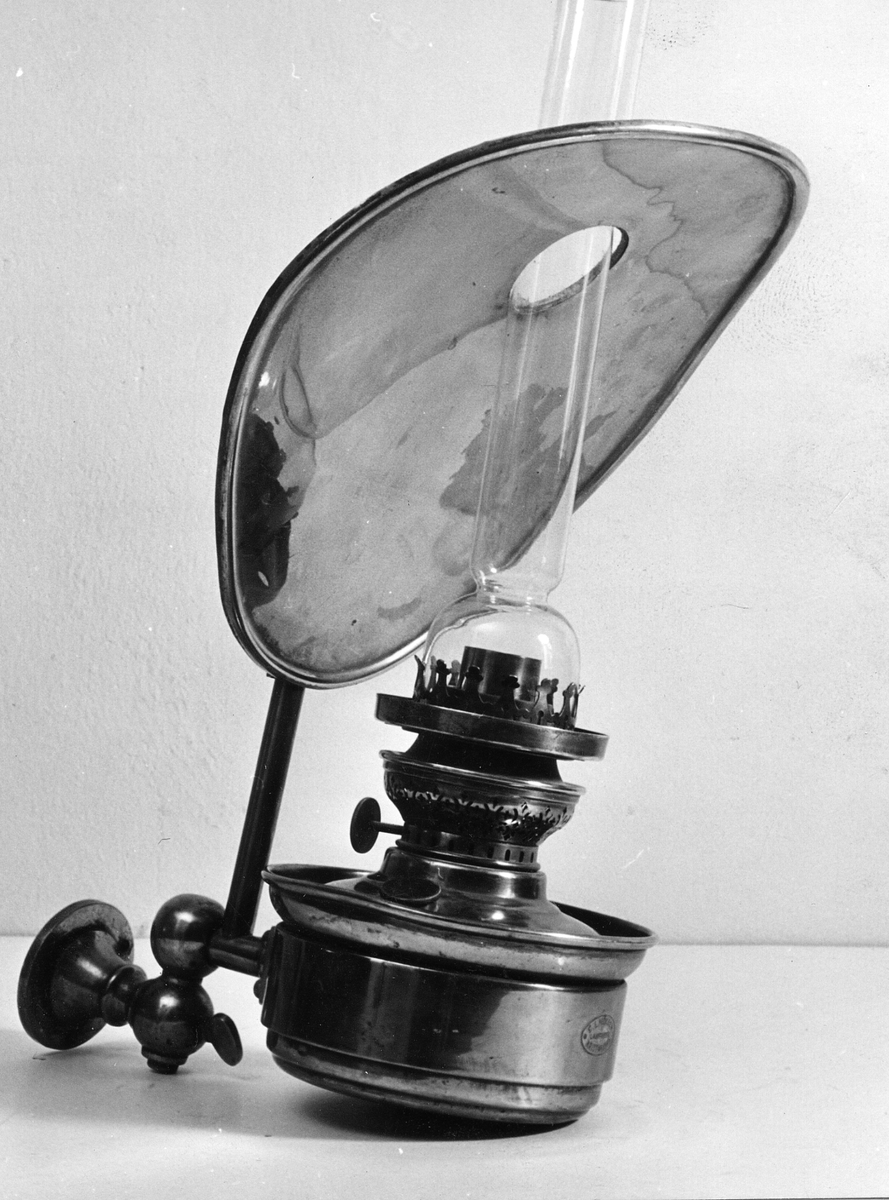 Fotogenlampa för postkupé. Av mässing med anordning för lampans fastskruvande vid vägg. Glaset går genom hål i snedställd hjärtformad reflexskiva.