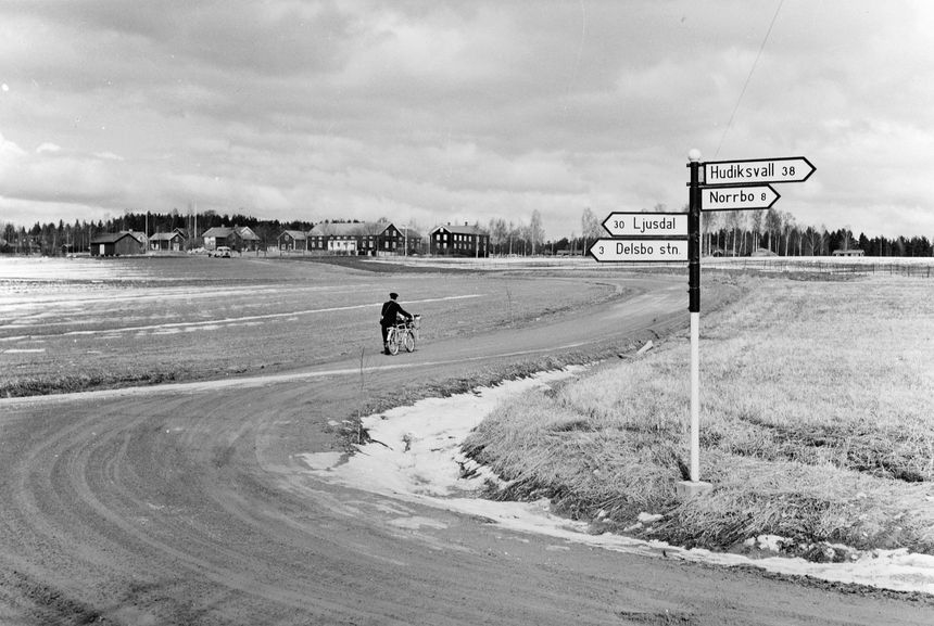 Lantbrevbärare, postiljon Erik Bergman (cykelåkande) i
Delsbo.  April 1956. Brevbäringsturen är 2 ½ mil lång och omfattar nästan 300 hushåll.  Postväskan väger ofta 30-40 kg.