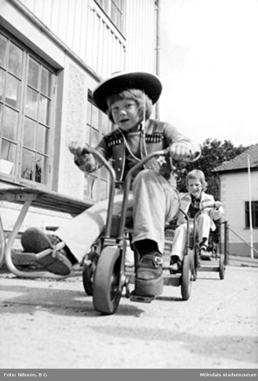 Två pojkar som cyklar på trehjulingar varav den främre är klädd i cowboy-kläder. Holtermanska daghemmet juni 1974.