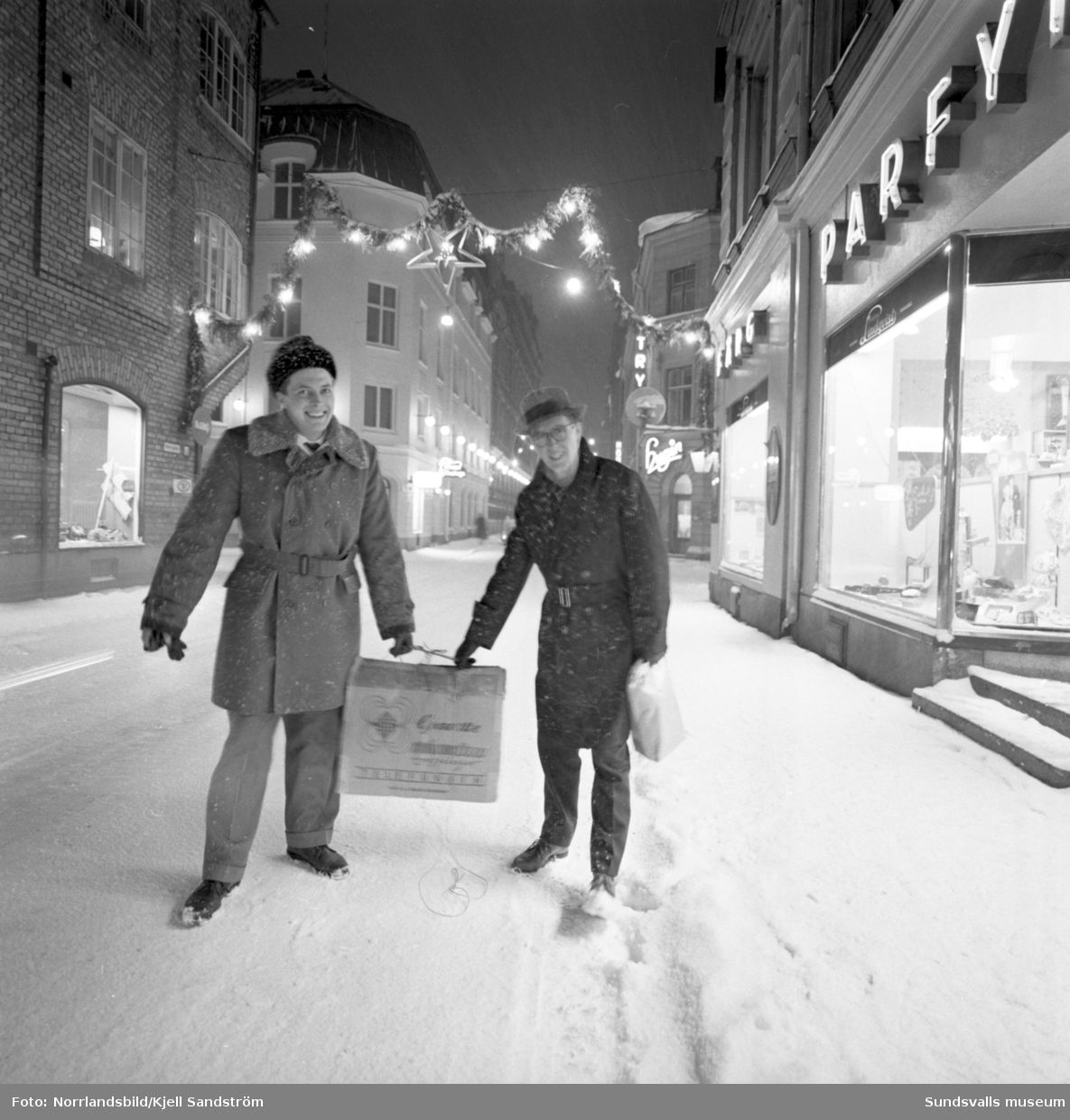 Kvällsbild från Nybrogatan i julbelysning. Två glada herrar bär hem sina julklappsinköp.