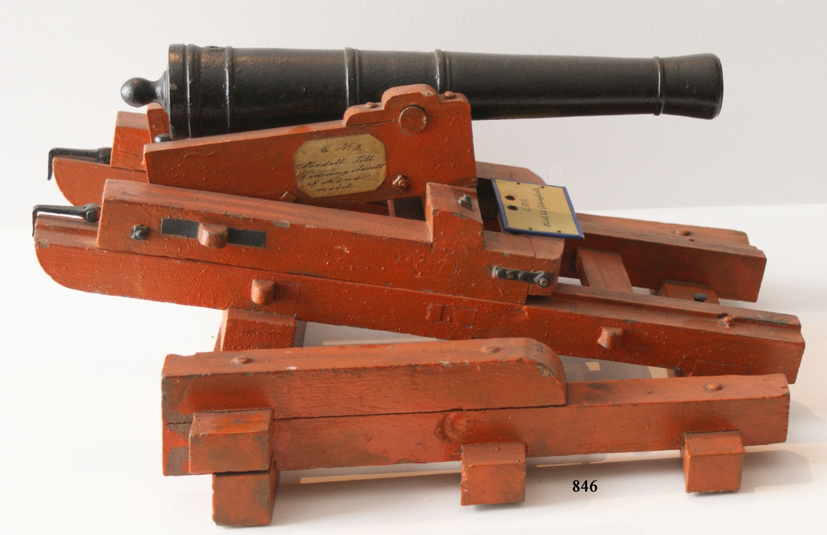 Kanonmodell: fästningslavett okänd modell med kanon av järn. Lavetten av trä, rödmålad. Lavetten L = 460 mm B = 440 mm H = 200 mm.