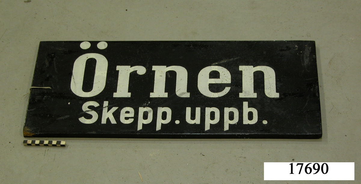 Rektangulär skylt, framsidan svartmålad, hål för upphängning. Vit schablonmålad text: " Örnen Skepp. uppbörd ".