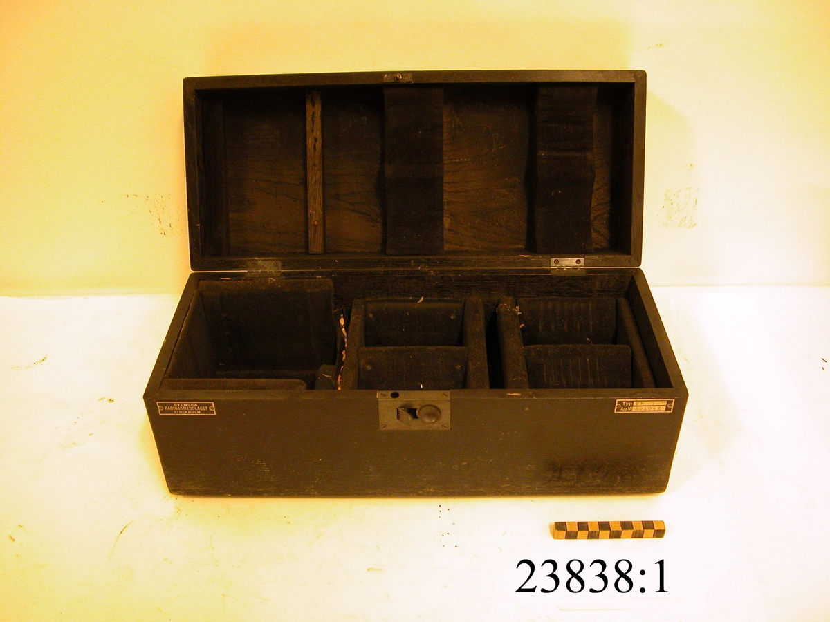Svart, rektangulär låda av trä med sex stycken fack fodrade med svart sammet. I lådan förvaras instrumentets fem komponenter MM 23838: 2-7