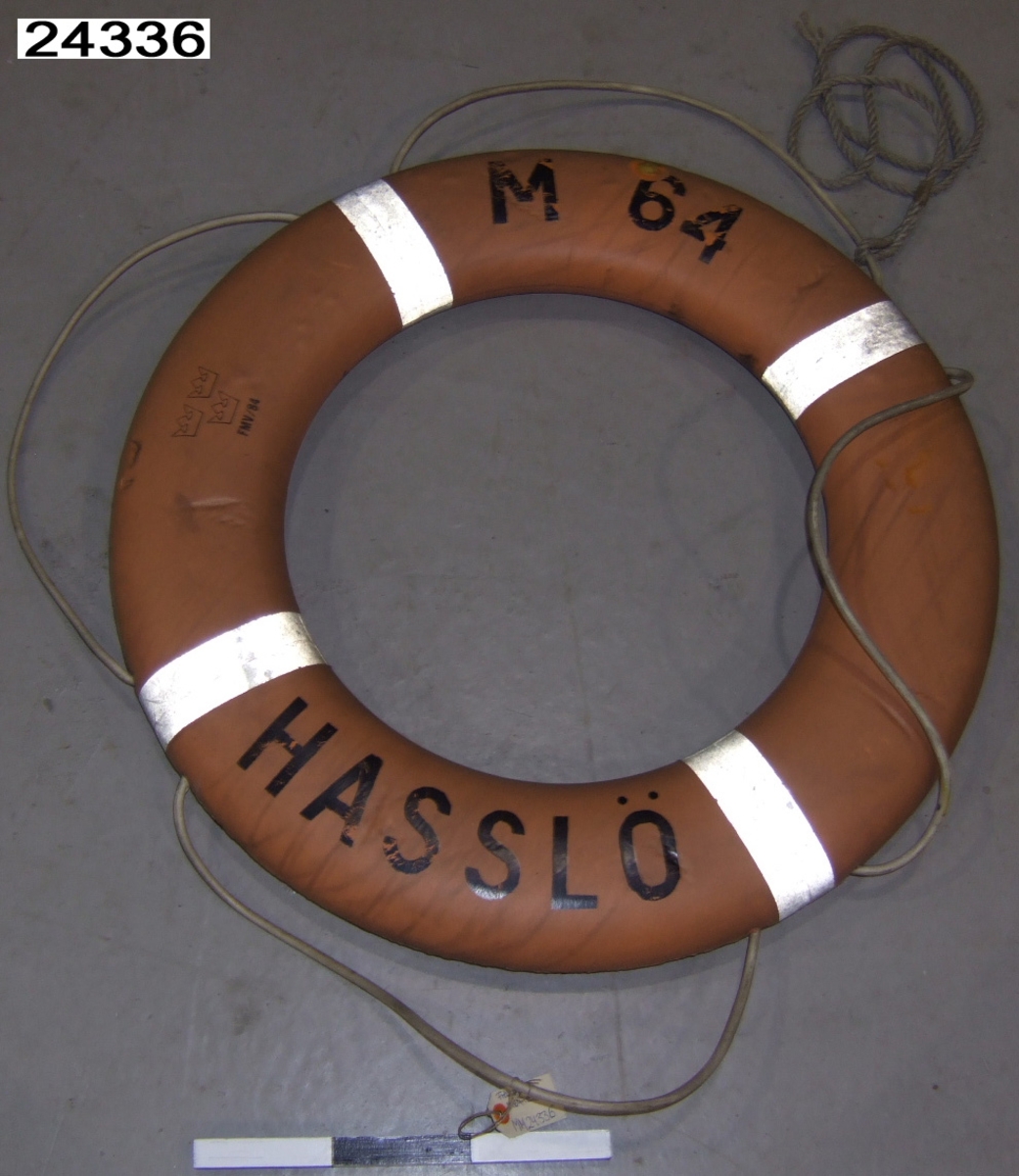 Orange frälsarkrans försedd med fyra reflexband och runtgående lina av gummi. Märkt "M 64, HASSLÖ", tre kronor och "FMV/84, M7383-204030, FRISTAD-PLAST AB, 2009/79" i svart.