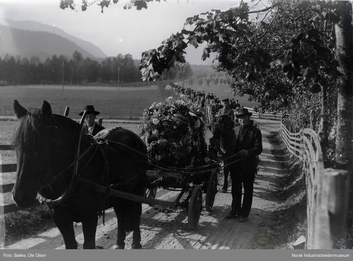 Stort gravfølge følger kiste som blir fraktet med hest og vogn langs landevei. Kisten på vognen er pyntet med båreblomster.