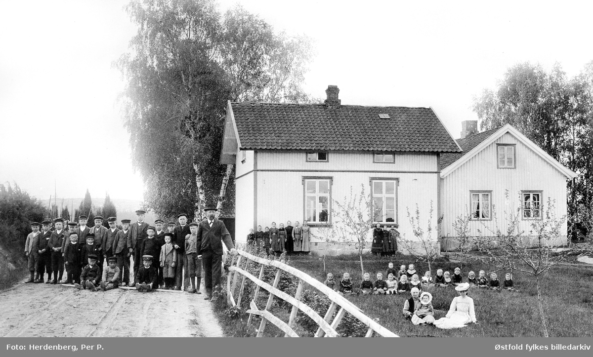 Nylende skole i Rolvsøy på slutten av 1890-tallet. Elevene er plassert i grupper; guttene til venstre på veien, de store jentene ved ny- bygget, de mindre jentene sitter i haven, og foran er lærerens familie. Lærerboligen ble skole og nybygg til lærer ferdig i 1892. Lærer Josef Olsen Hestenæs F.1853, kona Oline også lærer.