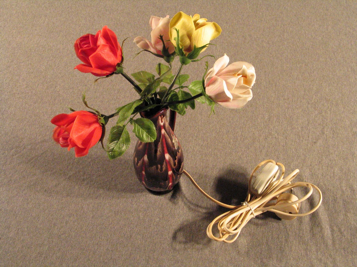 Lampe der foten er forma som ein vase med roser der det er sett små pærer i kvar av blomane. Slank konisk vase med hank og tut, oppi denne 2 raude, 2 rosa og 1 kvit plastrose.
Kvit plastledning med bryar midt på, truleg sekundær
