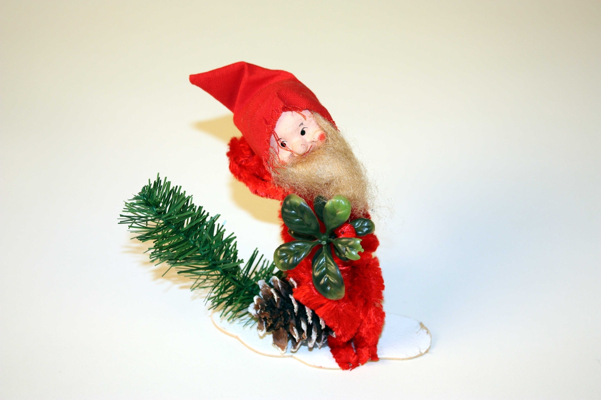 Julenisse av piperensar, sit på ei furukongle med snøglitter. Nissehovudet er av pappmache, måla ansikt, langt skjegg. Nissehue av stoff. Plastimitasjonar av granbar og tytebærlyng. 
Figuren er limt på ei papplate.