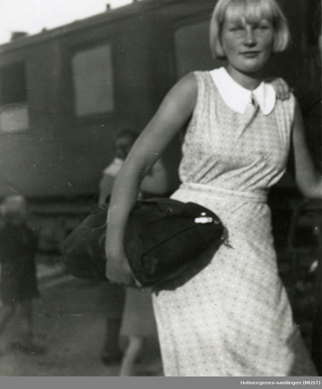 En ung kvinne med en veske under armen. En togvogn i bakgrunnen. Skrevet på baksiden: "Skoleturen til Nærland høsten 1933. Anne Marie Kvalsund. På Nærbø jernbanestasjon".