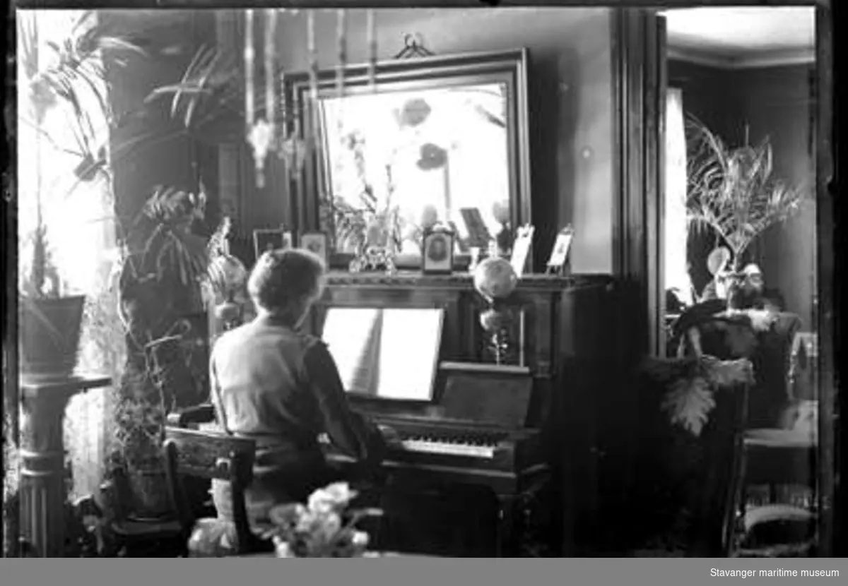 Borgerlig interiør. Kvinne som spiller piano ved et vindu. Pidestaller med bregnere.