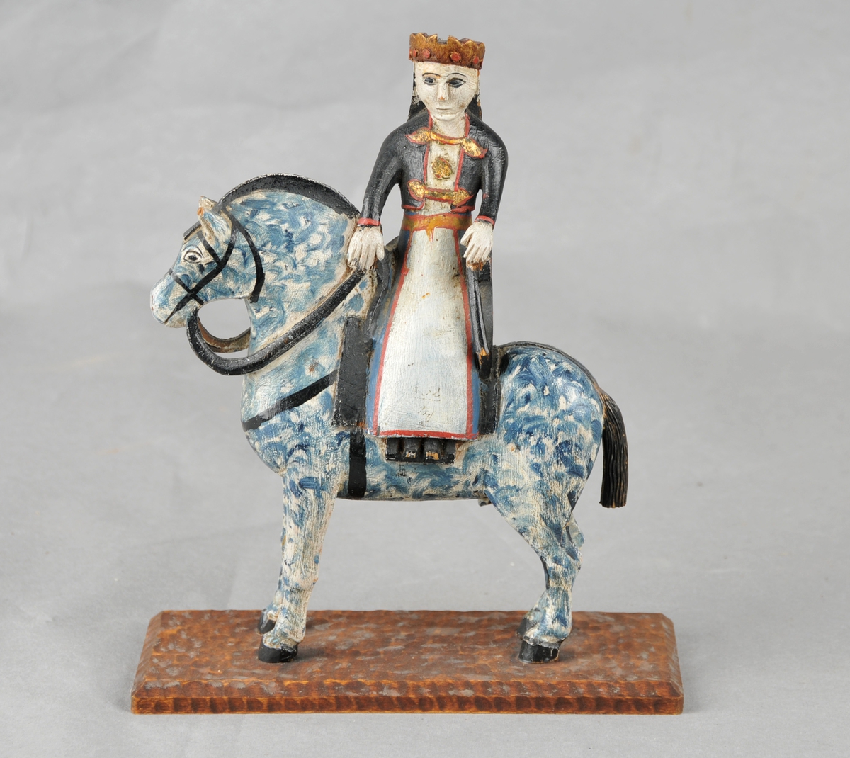 Liten skulptur utskoren i tre. Motivet er hest med brur. Brura sit i tverrsal, er pynta med krone på hovudet. 
Hesten er kvit med blå spettar. Heile figuren samt sokkelen er måla.