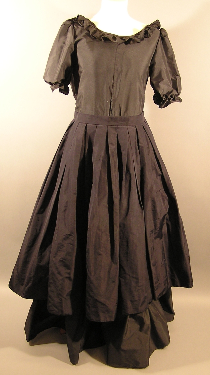 Gallakjole i to-deler. Enkel bluse med puff erme og rysje krave. Skjørt med bred linning, glidelås og hekte bak. Skjørt har tre lag. Kort skjørt øverst, underskjørt i midten og lang skjørt innerst. To sjal eller belter følger med. BKM.013423 og .013424.