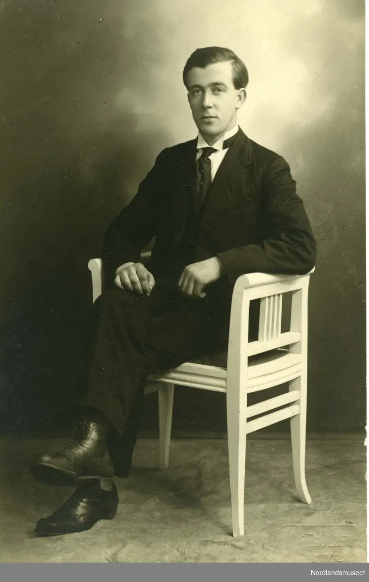 Portrettfoto fra postkort av en ung mann som sitter i en hvit trestol. Mannen har mørk dress, slips svarte sko og beina krysset over hverandre. Kortklipt hår.

Bakside: Ikke påskrevet.
