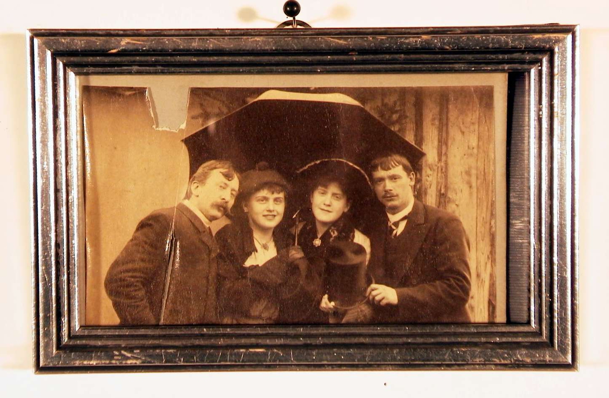 Fotografi av to unge damer i midten og en herre på hver side, samlet under en paraply.