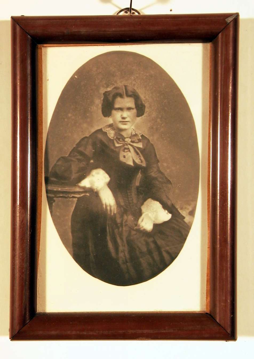 Portrettfotografi av en ung dame sittende med høyre arm hvilende på pidestalbord. Hun er ikledd en mørk kjole med snøreliv (korsett), store hvite kniplingsmansjetter og en stor sløyfe i halsen. Frisyren er oppsatt med midtskill.